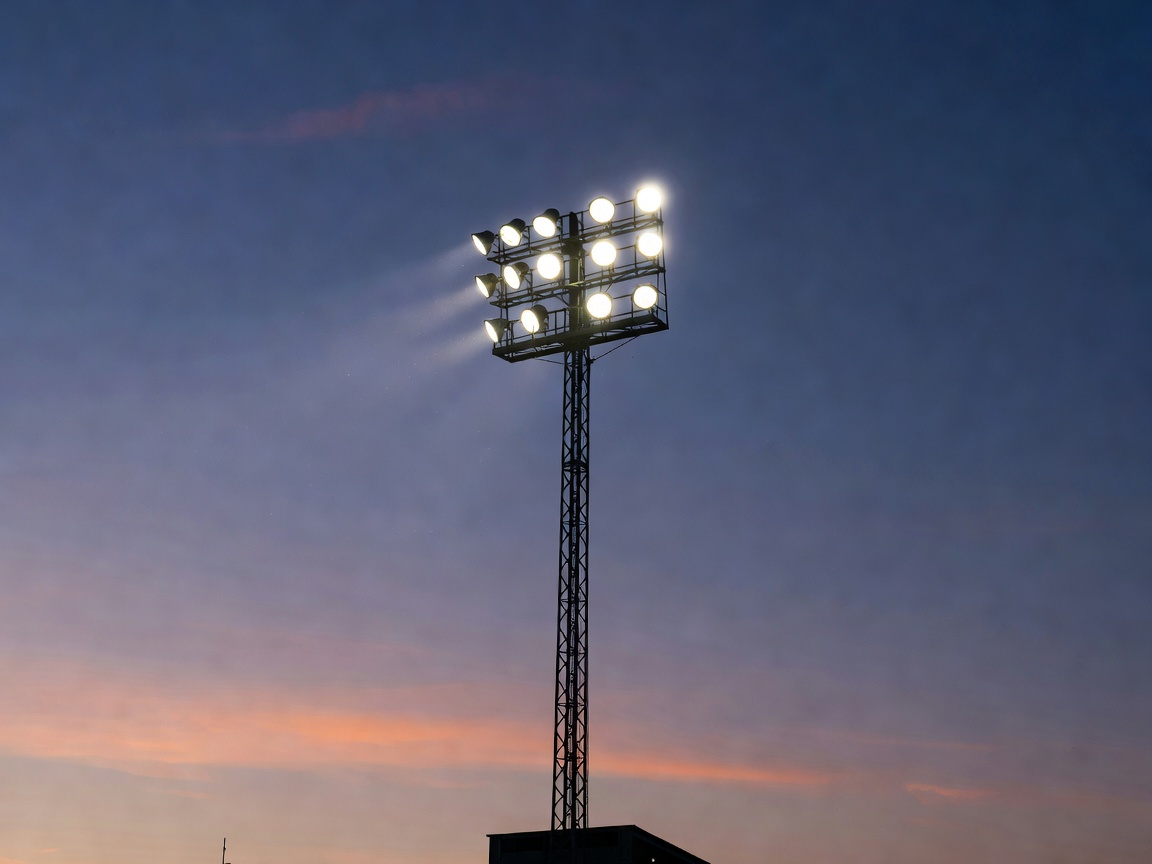 Tall stadium floodlight mast with multiple high-output projector lamps at dusk